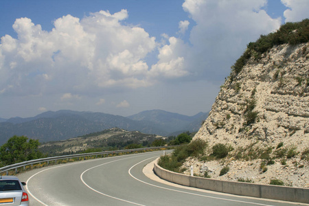 在一个阳光明媚的春天里，塞浦路斯的两条小巷柏油路的曲线上有一块白色的大石头，可以看到山脉和天空中的云的全景