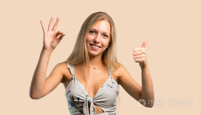 年轻的金发女人用手指显示一个OK标志，另一只手在Ocher背景上做一个拇指向上的手势