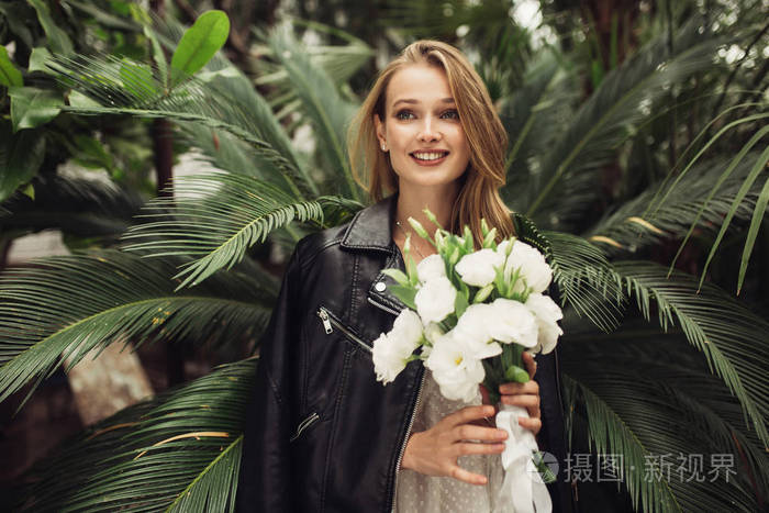 的女人穿着白色连衣裙,肩膀上穿着黑色皮夹克,手里拿着小新娘的花束