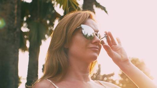 美丽时尚的金发碧眼的女人戴着太阳镜, 走在棕榈树的小路上。手掌反映在眼镜里