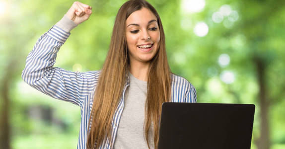 带手提电脑的带条纹衬衫的年轻女孩在户外庆祝胜利