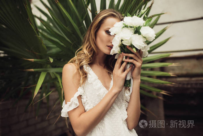 年轻漂亮的女人穿着白色的裙子,体贴地遮住脸,在舒适的温室里花着一束