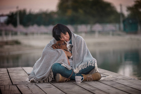 美丽的年轻女子拥抱她的狗在河边的肖像。 快乐的女人，带着她的狗贝塞特猎犬，裹着毯子，坐在夕阳下的木墩上