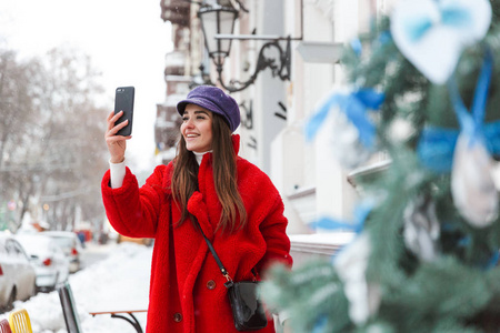 一个漂亮的年轻女人在户外走在雪冬街，用手机自拍。