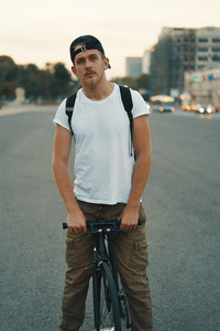 一个年轻人骑自行车在城市道路街道上的肖像，城市的背景很远。 男子骑黑色自行车，白色衬衫帽背包骑自行车去目的地