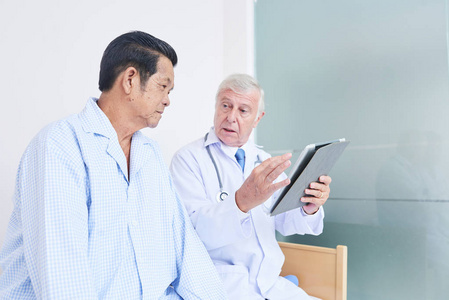 亚裔老人看现代平板电脑的侧视听医生坐在病房的床上
