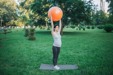 强壮的孕妇在公园里对瑜伽伙伴练习。她把橙色的大健身球放在头顶上
