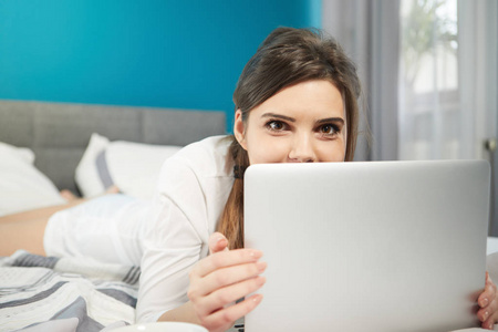年轻的女人在家里明亮舒适的卧室里躺在床上，手提电脑工作