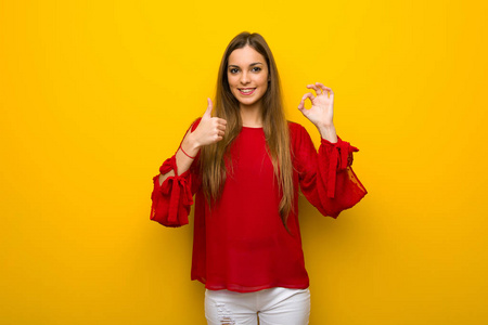 穿着红色裙子的年轻女孩穿在黄色的墙上，露出可以的标志，并伸出大拇指的手势