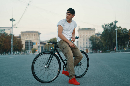 一个现代年轻人坐在街上骑自行车的户外肖像。 一个年轻的运动员穿着卡其裤白色T恤红色运动鞋和背包探索欧洲城市。