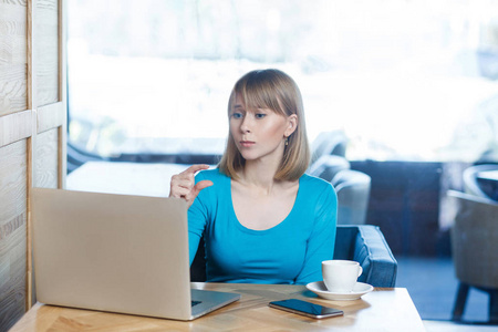漂亮的年轻女子，一头金发，穿着蓝色衬衫，坐在咖啡馆里，在笔记本电脑上打视频电话，同时让她的同伴更多地花在漂亮的脸上