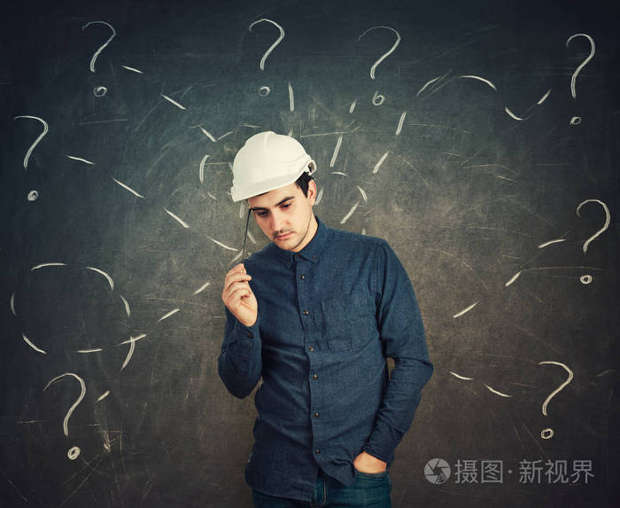 困惑的年轻人工程师戴着防护头盔,思考着用铅笔指向黑板背景上的头