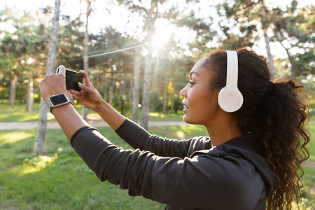 穿着黑色运动服和耳机的20多岁漂亮女人的肖像，在穿过绿色公园时，在手机上自拍照片。
