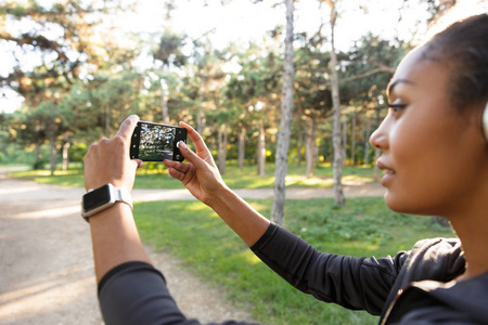 20多岁的年轻女子穿着黑色运动服和耳机，在穿过绿色公园时，在手机上拍照