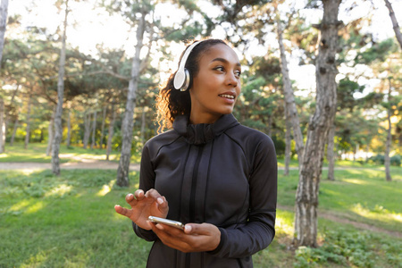 20多岁的运动女性穿着黑色运动服和耳机，在绿色公园散步时使用手机