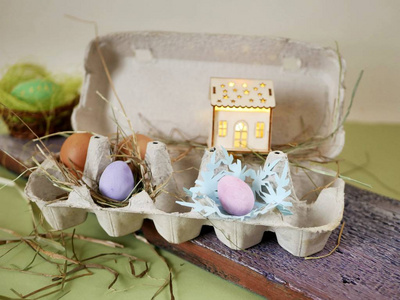 新鲜和复活节彩蛋鸡和鹌鹑蛋在包装托盘，一个玩具屋，照明在绿色的桌子上散落的干草，舒适的家庭假日复活节的概念。