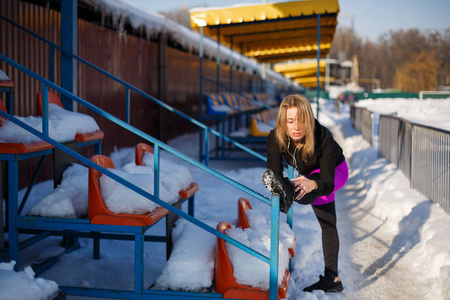 年轻的白种人女性金发碧眼，穿着紫罗兰色的紧身裤，在一个下雪的体育场的论坛上伸展运动。 健康和运动生活方式。