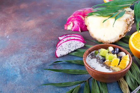 早餐冰沙碗，上面有火龙果，菠萝，辣椒，种子和浆果，棕榈叶在石头背景。