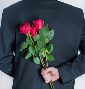 优雅的男人背着手捧红玫瑰花。 给女人的惊喜礼物。 浪漫和情人节假期的概念。 近距离选择性聚焦