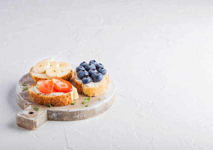 新鲜健康的迷你三明治与奶油奶酪水果和浆果在老式木板上。 草莓蓝莓香蕉和覆盆子在石制餐桌上的背景