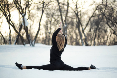 年轻的白种人女性金发碧眼，穿着紧身裤，在雪地森林里，坐在一根绳子上，伸展运动。 健康和运动生活方式。