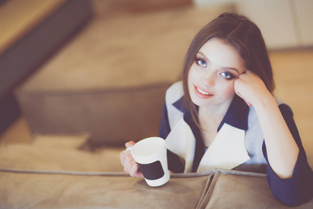 年轻的女人坐在舒适的沙发上，手里拿着杯子在白色的房间里喝咖啡。