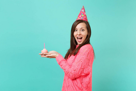 侧面观看兴奋的女人穿着针织粉红色毛衣，生日帽保持张嘴，看上去惊讶地拿着蛋糕与蜡烛隔离在蓝色背景上。人们的生活方式观念。把复制空间