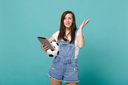 困惑的年轻女子足球迷拿着足球，使用平板电脑电脑，摊开双手孤立在蓝色绿松石墙背景上。人的情感运动家庭休闲生活理念
