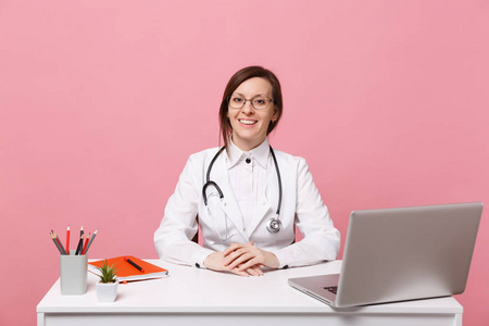 美丽的女医生坐在办公桌上，用电脑工作，医院里的医疗文件隔离在粉彩背景上。 穿着医用长袍眼镜的女人听诊器。 保健医学概念