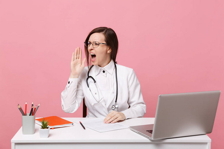 疲惫悲伤的女医生坐在电脑上的办公桌上工作，医院里的医疗文件孤立在粉墙背景上。 穿着医用长袍眼镜的女人听诊器。 保健医学概念
