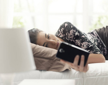 年轻漂亮的女人在床上放松，并与她的智能手机连接，她正在和她的朋友聊天，阅读信息