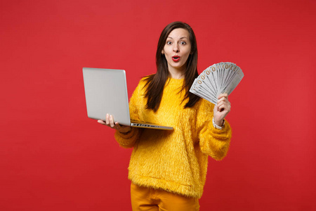 惊讶的年轻女子穿着黄色毛皮毛衣，手提电脑电脑风扇的钱，美元，现金，孤立在红色背景。 人们情感生活方式的概念。 模拟复制空间