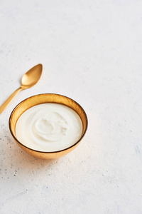一碗新鲜的希腊酸奶和金色勺子在白色抽象的背景上。 健康的早餐。 黄金器皿。 上面的风景。 复制文本空间。