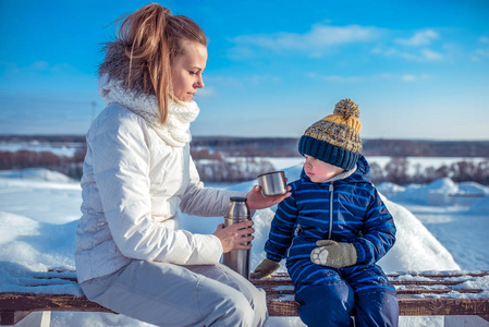 年轻的母亲从热水瓶里倒了一杯茶。小男孩的孩子看着杯子。冬天, 在城市里, 在一个雪院的长凳上