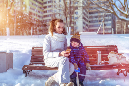 年轻的母亲向孩子展示智能手机。冬天, 他们坐在长凳上玩, 在街上的一个城市公园里玩。快乐的微笑。穿着休闲装附近有茶和包的热水瓶