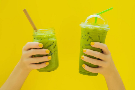 绿茶拿铁与冰在马松罐和在一个塑料杯在手在黄色背景。自制的冰抹茶拿茶与牛奶零浪费