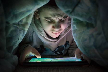 孩子在床上看着他的平板电脑。 从设备屏幕上照亮儿童脸。 被子下面的男孩拿着一块平板电脑。 晚上的时间。