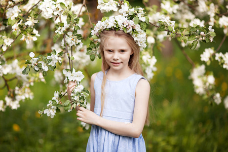 美丽的年轻女孩穿着蓝色的裙子在花园里，有着耀眼的苹果树。 可爱的女孩抱着苹果树枝