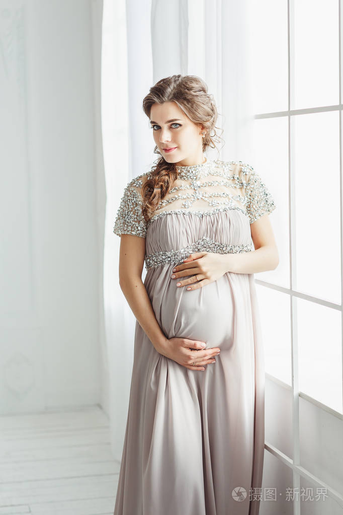 一位优雅美丽的孕妇的肖像,穿着灰色的连衣裙,在白色的工作室里摆姿势