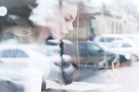 一个有吸引力的年轻记者女孩的肖像与笔和记事本在咖啡馆背后的展示。通过窗户玻璃反射的非对比度视图