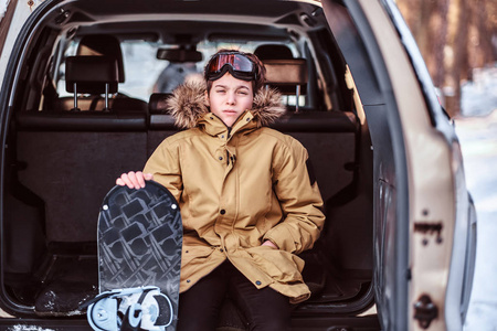 一名少年滑雪者穿着暖和的衣服坐在一辆汽车的后备箱上