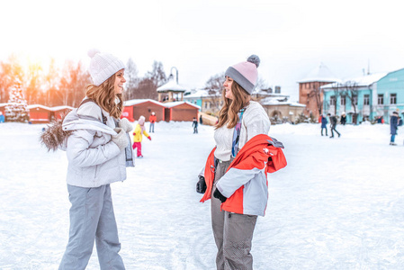 两个美丽的女孩女友, 妇女姐妹快乐微笑着在城市的冬天说话。穿着白色保暖的休闲服和帽子。在冬日下午休息, 背景是人们在溜冰场上溜冰