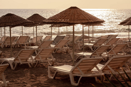 地中海海滩海岸线海景，傍晚日落时有草遮阳和舒适的白色甲板椅