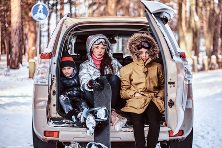 一个漂亮的女人和她的儿子们穿着温暖的衣服坐在汽车后备箱上