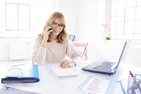 中年休闲女商人坐在笔记本电脑前的办公桌前工作时,用手机与某人交谈