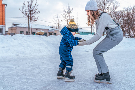 一位年轻的母亲牵着一个24 小男孩的手。冬天, 在城市里溜冰场上。您可以在新鲜的冷风中放松身心。溜冰鞋的第一步, 家长的帮助和