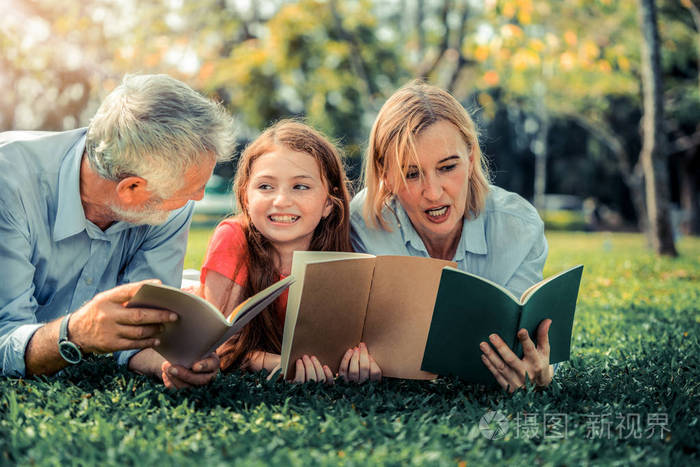 幸福的家庭一起看书躺在公共公园的绿草上小女孩和父母一起在户外花园