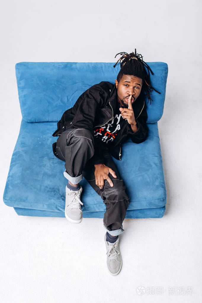 一个迷人的微笑的黑人穿着黑色夹克,穿着灰色背景的黑裤子,坐在蓝色