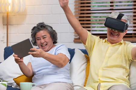 亚洲资深夫妇玩虚拟现实眼镜耳机和平板电脑，观看VR视频，并在家客厅的沙发上玩得开心。