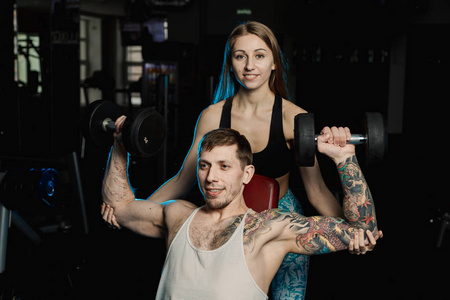 活跃美丽的健身模型女孩帮助训练与哑铃, 而坐在她的男朋友在健身房